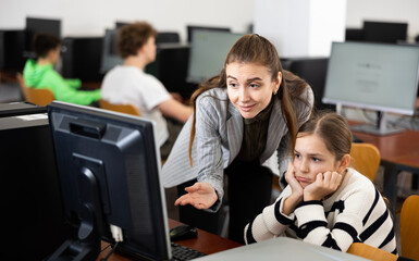 Caring young female teacher calming upset teenage schoolgirl sitting at computer in classroom..