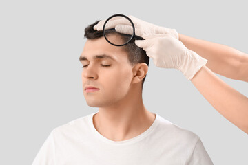 Doctor with magnifier examining young man's hair on light background, closeup