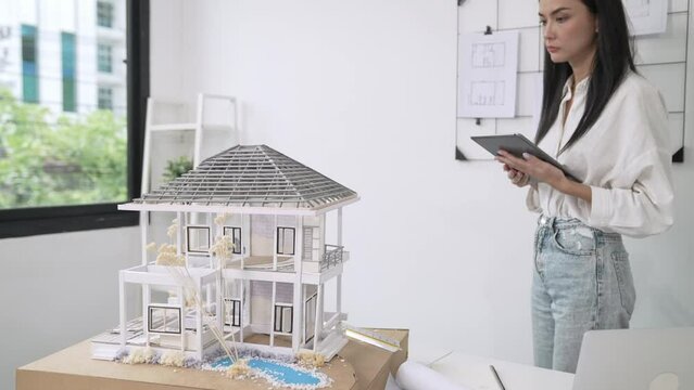 Beautiful caucasian architect focus on using tablet to check house model on meeting table with blueprint, house model and architectural document scatter around at meeting room. Immaculate.