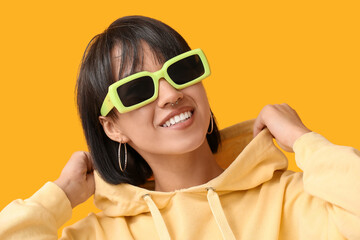 Happy stylish young woman in sunglasses with nose piercing on yellow background