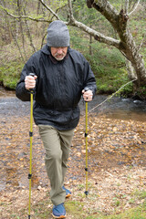 Senior hiker in cold weather gear crossing a creek using trekking poles 