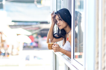 A woman in a summer dress holds a mug in an open window of a residential building as the wind blows through her hair - obrazy, fototapety, plakaty