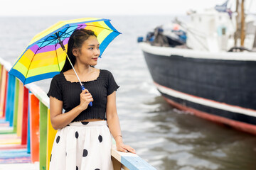 A young woman with a parasol stands on a colorful footbridge and watches a sailing ship