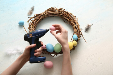 Woman making Easter wreath with  painted eggs and glue gun on color background