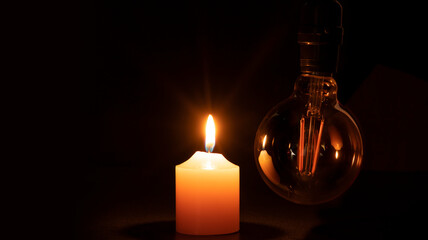 Blackout concept, turned off light bulb with no electricity hanging next to a burning candle - obrazy, fototapety, plakaty