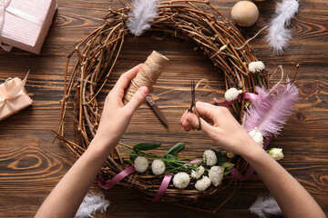 Woman making Easter wreath with eggs, feathers and flowers on brown wooden background