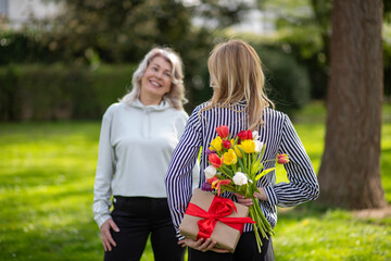 Daughter surprises mother with hidden gift and flowers behind back.