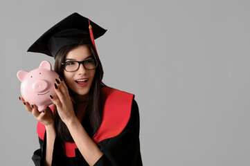 Beautiful young happy female graduate with piggy bank on grey background. Tuition fees concept