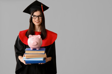Beautiful young happy female graduate with piggy bank and books on grey background. Tuition fees...
