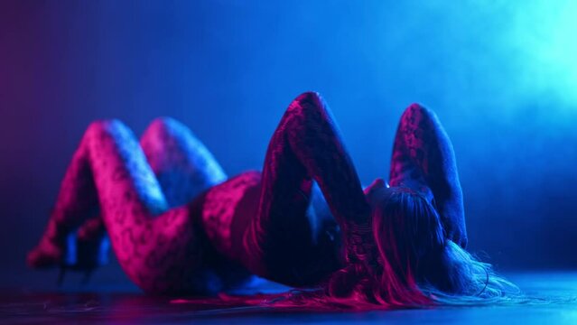 Sexy woman performs flexible movements with body on floor. Erotic hot dance in dark studio with neon light. Lady dancer in tight clothes and heels.