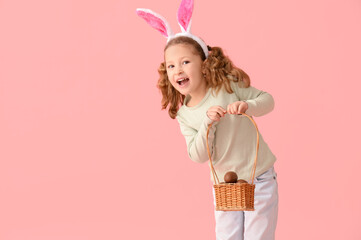 Cute little girl in bunny ears with basket of chocolate Easter eggs on pink background