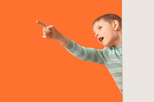 Shocked little boy with blank poster pointing at something on orange background
