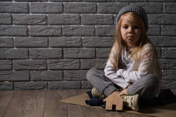 Homeless little girl with figure of house sitting near grey brick wall