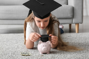 Little girl putting mortar board on piggy bank at home. Education savings concept