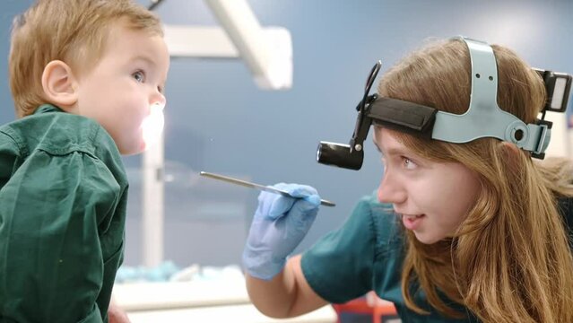 A cute toddler boy with his mother are at an appointment with a pediatric ENT. The doctor otolaryngologist is examines the throat of a little patient with inspection spatula