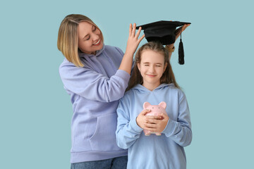 Mother putting mortar board on her daughter with piggy bank on blue background. Education savings...