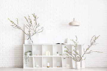 Vases with blooming branches on shelving unit near white brick wall
