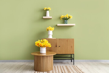 Vases with yellow narcissus flowers on coffee table and chest of drawers in green room