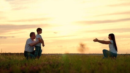 Child Boy runs from father to mother on meadow in evening. Kid with parents spends time together on field at sunset twilight. Boy runs from father to mother reaching out arms in embrace. Happy family - obrazy, fototapety, plakaty