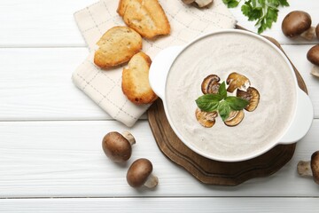 Delicious homemade mushroom soup in ceramic pot, croutons, fresh champignons and parsley on white wooden table, flat lay