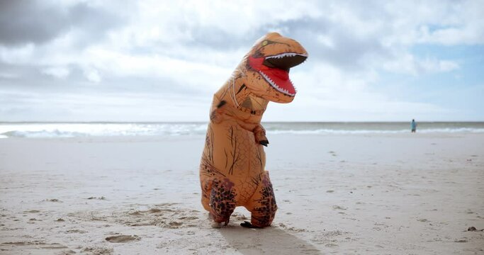 Animal, funny and dinosaur costume on beach dancing with energy for comedy joke on vacation travel. Goofy, silly and person with inflatable mascot moving, jumping and having fun by ocean on holiday.
