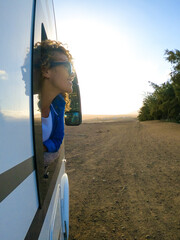 Blonde woman with curly hair looks out of the window of the moving camper. Female tastes the wind in her face and smiles. Travel and tourism concept.