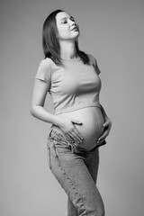 Black and white portrait of young pretty pregnant woman in t-shirt and jeans on gray background. - 787710455