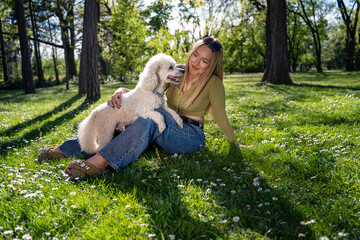 Young beautiful woman with her pet poodle sitting on the grass in the park, on beautiful sunny spring day 