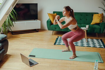 Sporty woman squats on mat in front of laptop in living room. Home workout concept