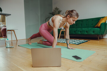 Woman in sportswear kneeling on mat in front of laptop on hardwood flooring