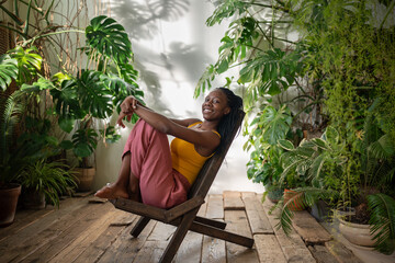 Joyful happy african american woman rest on chair surround by tropical houseplants enjoy calmness in sunny day look at camera. Playful pleased black young female smiling sit in room with indoor plants