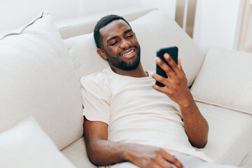 Happy African American man sitting on a black sofa, chatting on his smartphone He is dressed in a...
