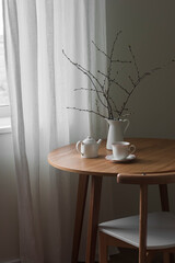A cup of tea, a teapot, branches in a jug on a round wooden table in the living room - 787772041