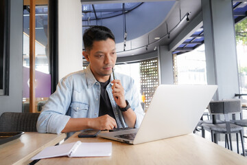 Asian man think and look for ideas with blue shirt using laptop and mobile phone in coffee shop,...