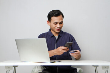 joyfull happy asian man holding mobile phone and credit card while sitting in front of laptop computer. on isolated background