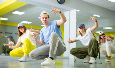 Expressive teen boy and three teenagers in caps sitting in breakdance pose in dance hall