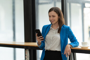 Young businesswoman smiling, holding a smartphone and leaning on a counter, enjoying a coffee break in a modern office environment, connecting with technology and staying productive