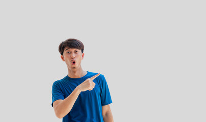 A young Asian man in his 20s wearing a blue t-shirt happy shocked face pointing thumbs up isolated on a gray background. The person's index finger pointing to a blank space for text