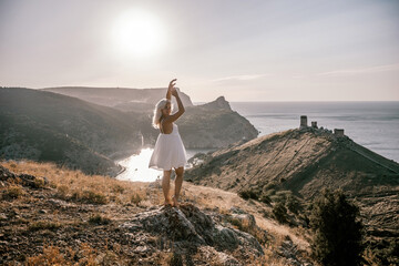 A woman stands on a hill overlooking a body of water. She is wearing a white dress and she is...