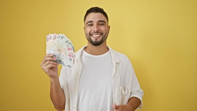 Cheerful young arab man flashing uae banknotes and joyful thumbs up, radiating confidence over yellow isolated background