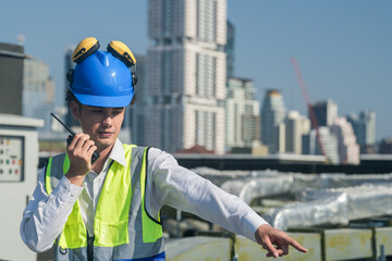 Focused engineer in hard hat and safety vest using walkie-talkie and laptop on construction site...