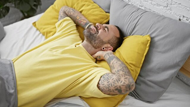 A young tattooed hispanic man with a beard, dressed in a yellow shirt, lying in pain on his bed while holding his neck.