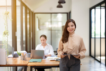 Confident businesswoman holding a clipboard, ready to present in a bright modern office, with colleague working in the background.