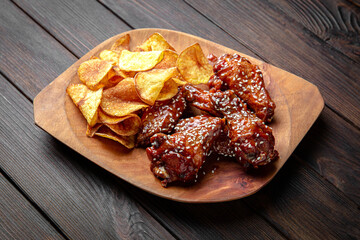 Spicy BBQ chicken wings with sesame seeds and potato chips on dark boards background. Menu for a pub