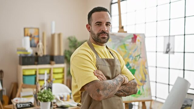 A confident tattooed hispanic man wearing a yellow shirt and apron stands arms crossed in a bright art studio.