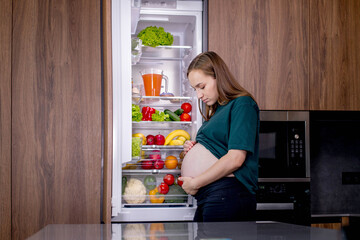 Hungry pregnant woman standing near refrigerator looking for food during pregnancy. Healthy eating