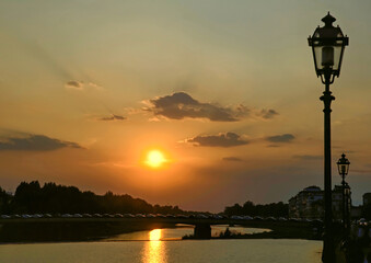 Orange, evening sunset on the Arno River in Florence, Tuscany, Italy.  Silhouetted lamp posts, clouds and bridge. Orange reflection on water. No people. - obrazy, fototapety, plakaty