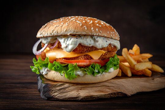 Fish burger with tartar sauce, cheese, vegetables, gravy and iceberg lettuce. Juicy delicious hamburger on darkmood picture for restaurant decoration, poster. 
