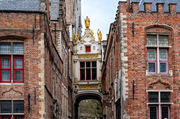 White bridge over a street in Brugge, with gold sculptures and red windows.
