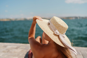 A woman in a swimsuit sits with her back holding a hat, looks at the ocean, sunny day, relaxes.